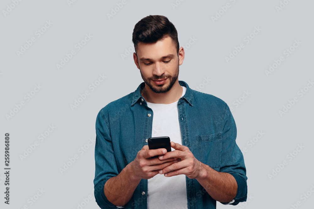 Texting to friend. Charming young man using his smart phone and smiling while standing against grey background Texting to friend. Charming young man using his smart phone and smiling while standing against grey background
