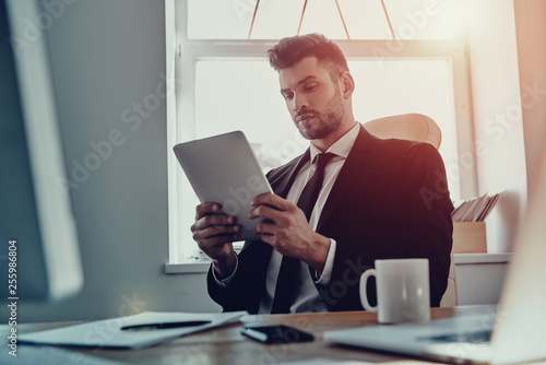 Businessman at work. Thoughtful young man in formalwear using digital tablet while sitting in the office