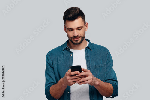Texting to friend. Charming young man using his smart phone and smiling while standing against grey background