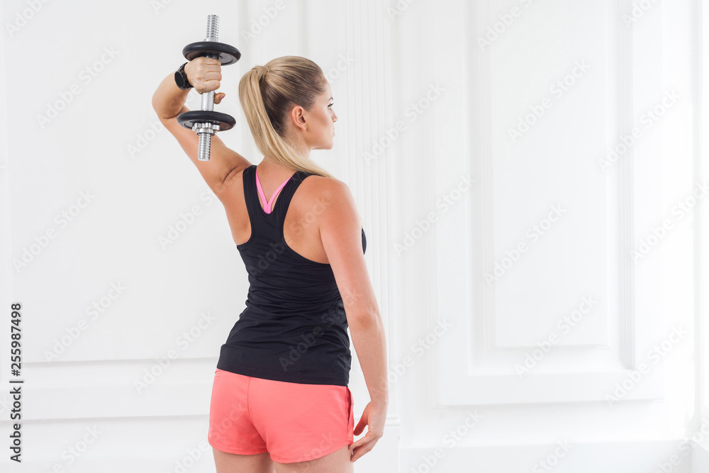 Striving for strong. Backside of young adult motivation athletic bodybuilder woman in pink shorts and black top standing, holding dumbells doing exercise for arms at the gym with perfect figure. Striving for strong. Backside of young adult motivation athletic bodybuilder woman in pink shorts and black top standing, holding dumbells doing exercise for arms at the gym with perfect figure.