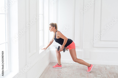 Side view portrait of young athletic beautiful bodybuilder woman in pink shorts and black top holding dumbells doing exercise for arms at the gym, looking at the window. indoor, studio shot, sport