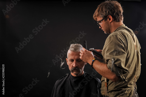 Hairdresser cuts senior citizen with a beard on a dark background in the studio