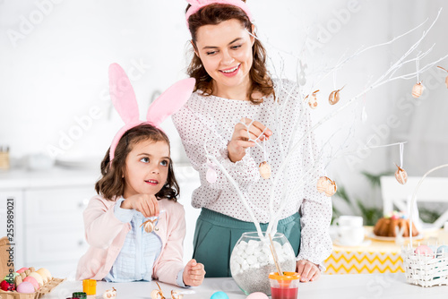 Smiling mother and daughter in bunny ears decorating easter tree in kitchen