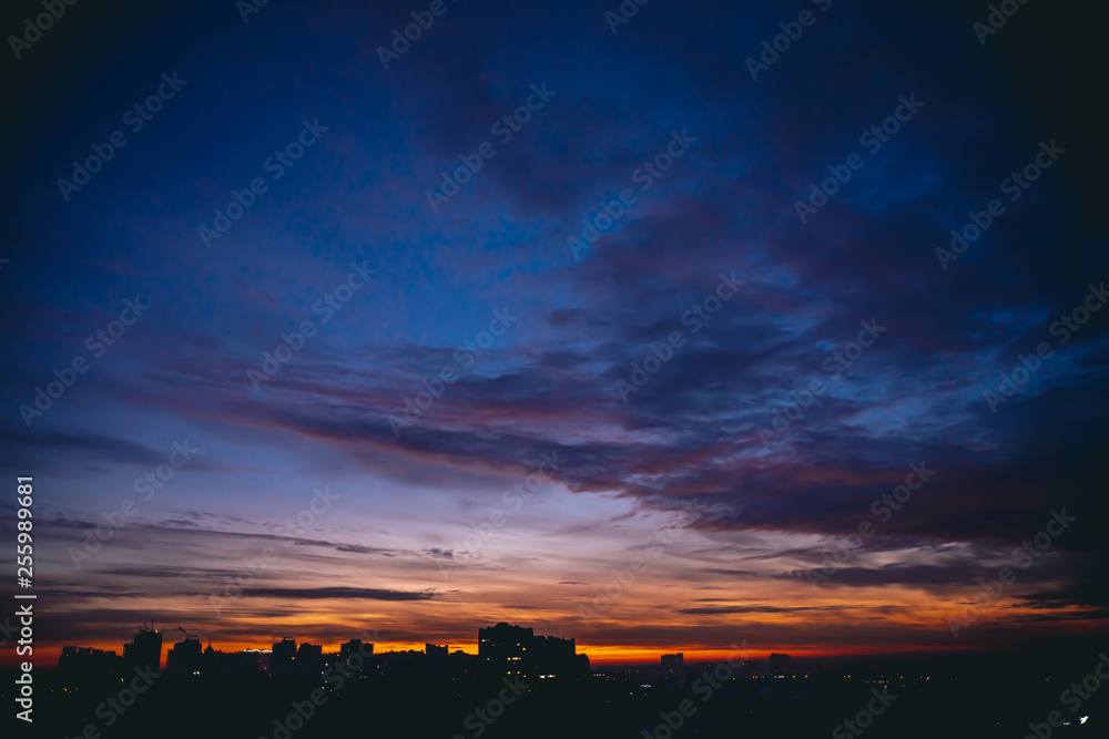 Cityscape with vivid warm dawn. Amazing dramatic blue violet cloudy sky above dark silhouettes of city buildings. Orange sunlight. Atmospheric background of sunrise in overcast weather. Copy space.