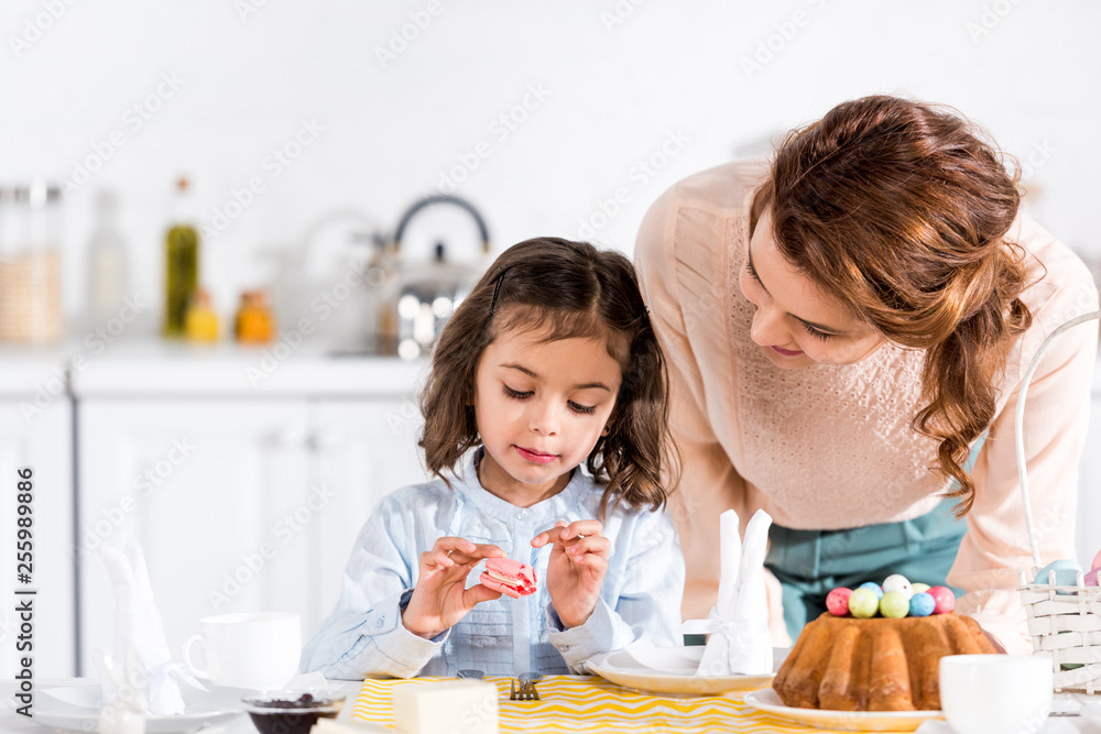Brunette woman looking at daughter eating macaroon in kitchen Brunette woman looking at daughter eating macaroon in kitchen