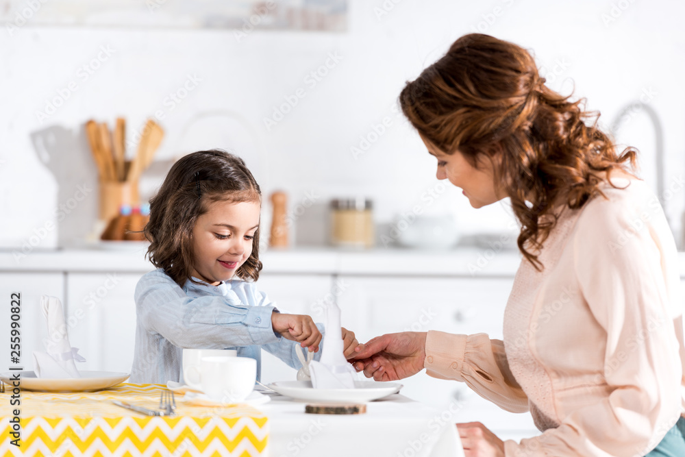 Brunette mother and daughter folding napkins at table in kitchen