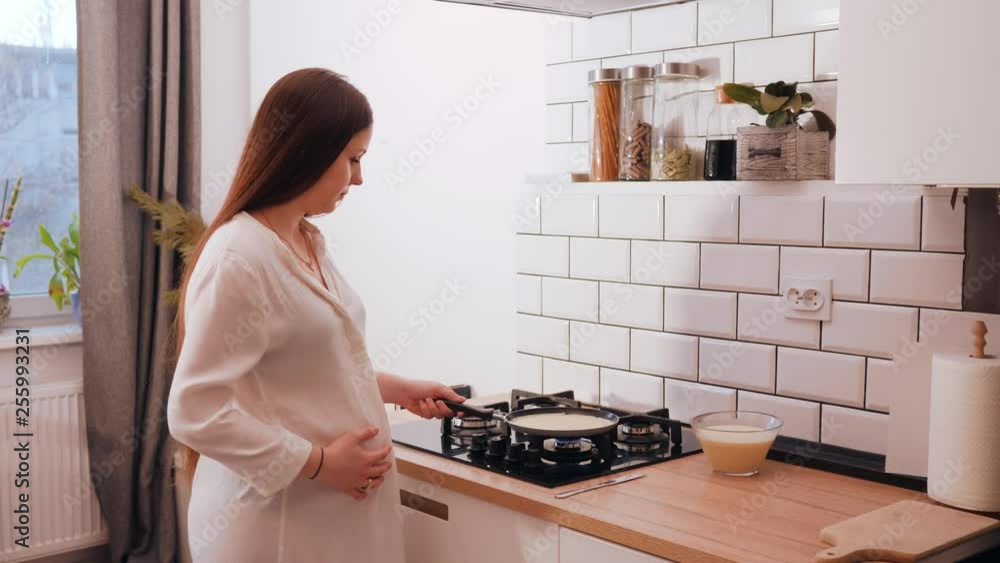 Young pregnant housewife cooking pancakes standing in the kitchen at the stove Young pregnant housewife cooking pancakes standing in the kitchen at the stove