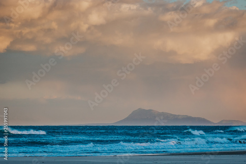 Beach scenery with blue waves, dramatic sky and mountains in the background. © Darius SUL