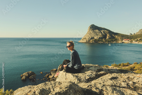 profile portrait of young stylish girl with hair bun and sunglasses is sitting on the rocky precipice, edge of mountain, blue sea below
