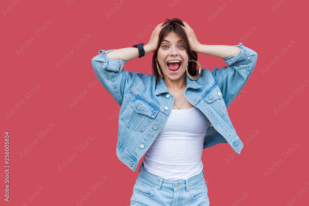 Portrait of surprised beautiful brunette young woman with makeup in denim casual style standing, holding her head, looking at camera with shocked face. indoor studio shot, isolated on red background. Portrait of surprised beautiful brunette young woman with makeup in denim casual style standing, holding her head, looking at camera with shocked face. indoor studio shot, isolated on red background.