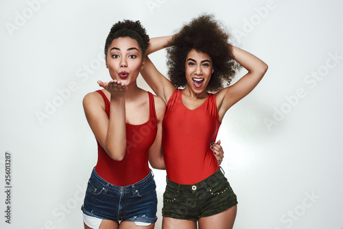 Feeling happy! Cute afro american woman in casual clothing sending an air kiss while while standing with her best friend against grey background