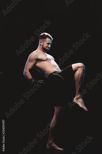 muscular barefoot strenuous boxer in boxing gloves doing kick isolated on black