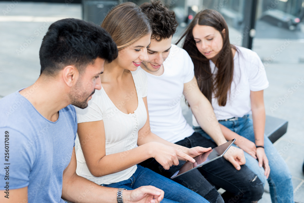 Group of young friends using a tablet together while sitting on a bench in a modern city Group of young friends using a tablet together while sitting on a bench in a modern city