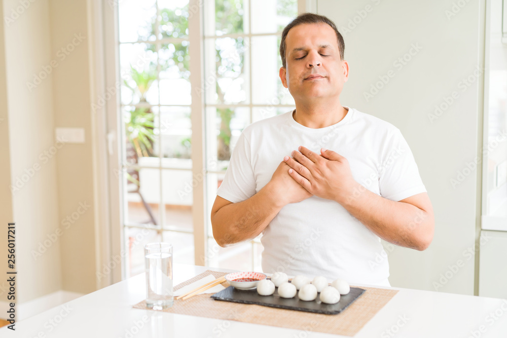 Middle age man eating asian dumplings at home smiling with hands on chest with closed eyes and grateful gesture on face. Health concept. Middle age man eating asian dumplings at home smiling with hands on chest with closed eyes and grateful gesture on face. Health concept.