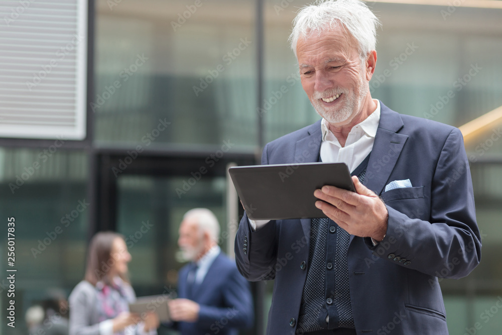 Smiling senior gray hair businessman using a tablet, standing on a sidewalk in front of an office building. Two people talking in the background, blurred Smiling senior gray hair businessman using a tablet, standing on a sidewalk in front of an office building. Two people talking in the background, blurred