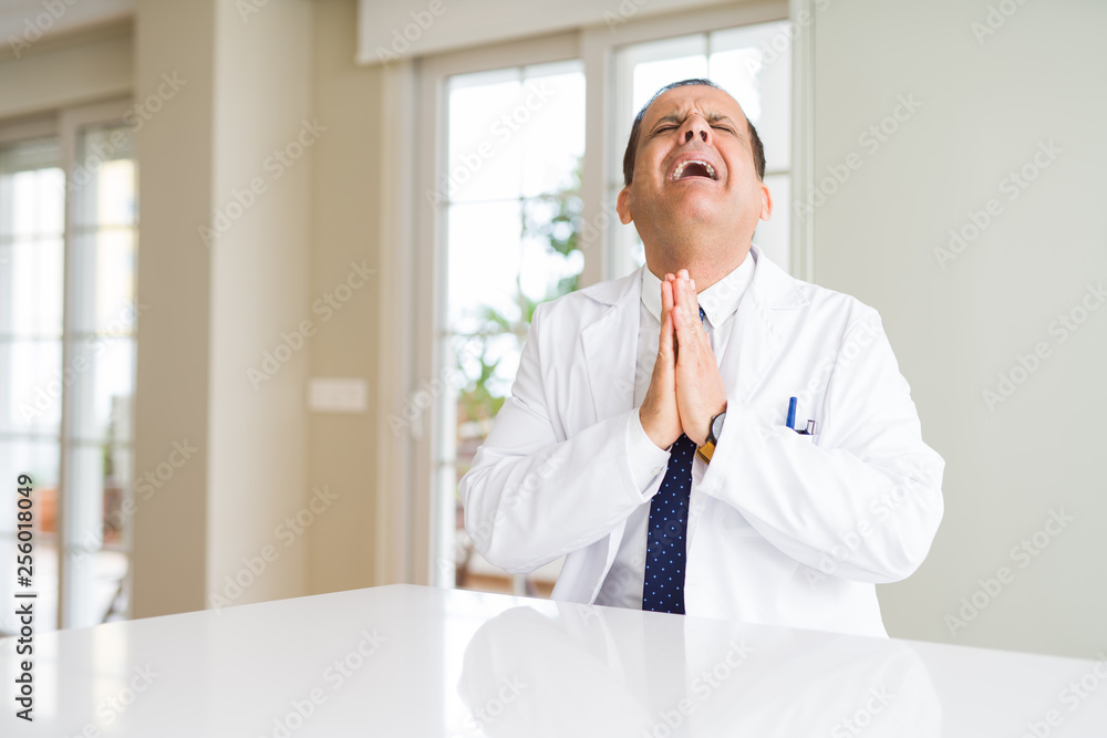 Middle age doctor man wearing medical coat at the clinic begging and praying with hands together with hope expression on face very emotional and worried. Asking for forgiveness. Religion concept.