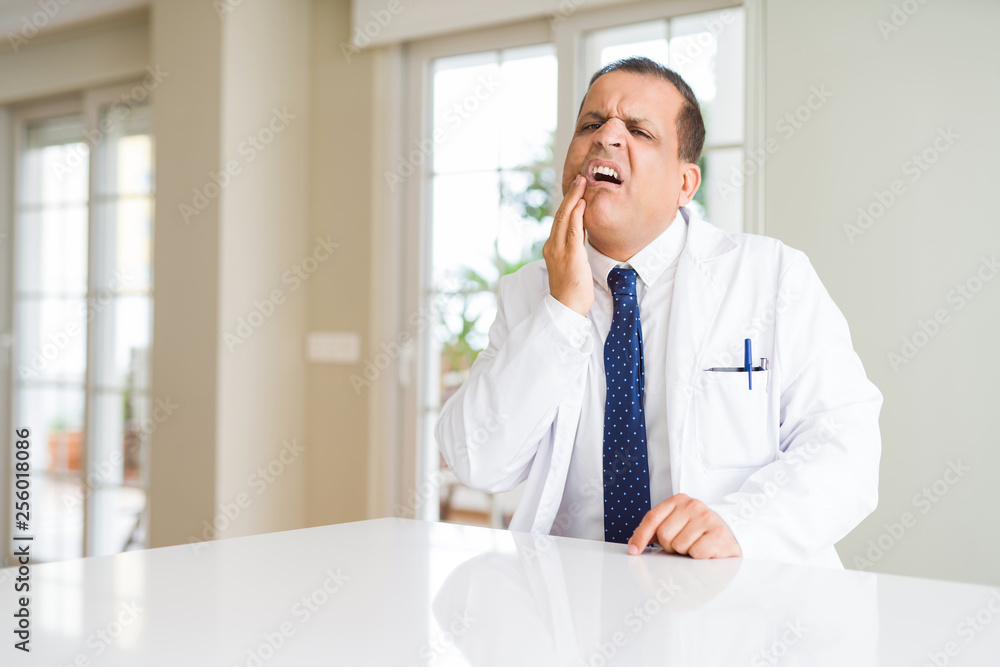 Middle age doctor man wearing medical coat at the clinic touching mouth with hand with painful expression because of toothache or dental illness on teeth. Dentist concept.