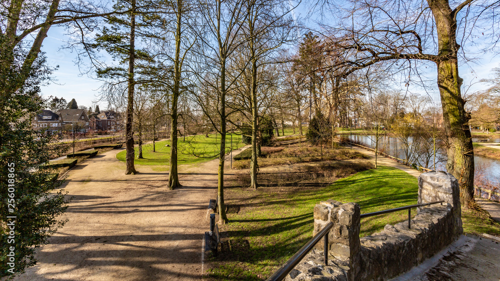 Landscape of Proosdij Park with its paths between bare trees, small river, against blue sky, view from stone bridge, sunny winter day in Meerssen, South Limburg, The Netherlands Landscape of Proosdij Park with its paths between bare trees, small river, against blue sky, view from stone bridge, sunny winter day in Meerssen, South Limburg, The Netherlands