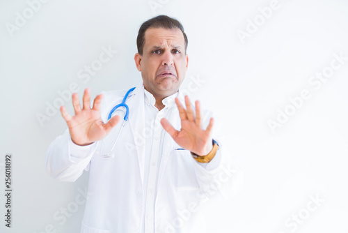 Middle age doctor man wearing stethoscope and medical coat over white background afraid and terrified with fear expression stop gesture with hands, shouting in shock. Panic concept. © Krakenimages.com