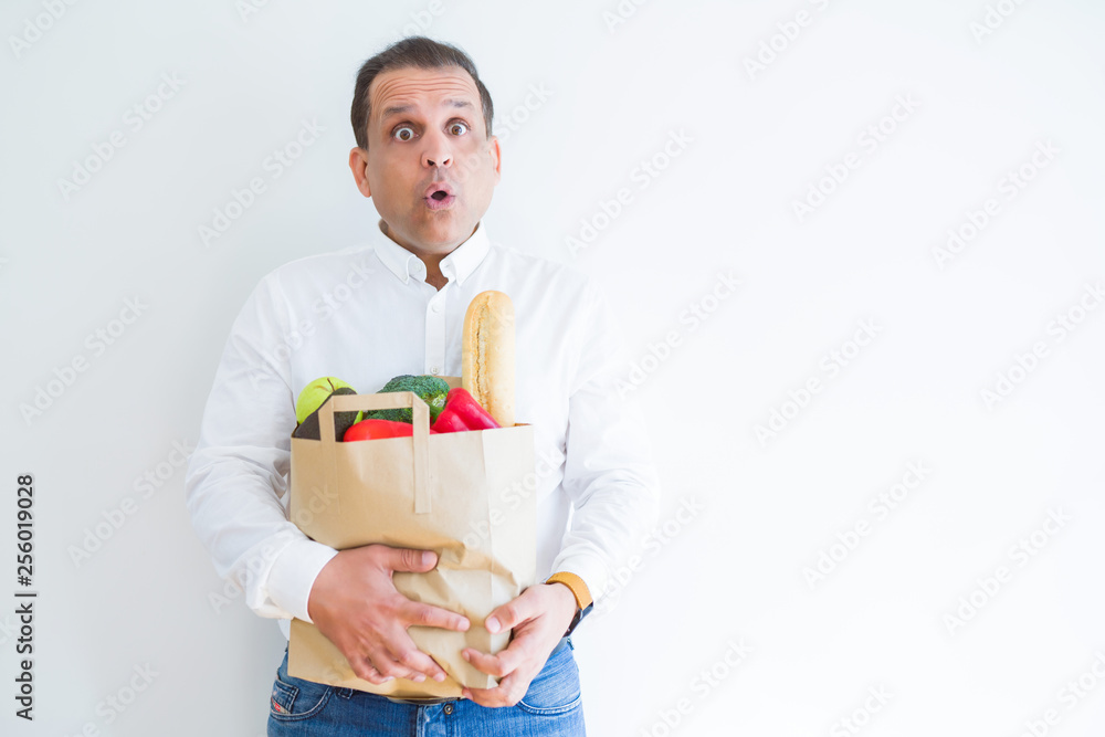 Middle age man holding groceries shopping bag over white background scared in shock with a surprise face, afraid and excited with fear expression Middle age man holding groceries shopping bag over white background scared in shock with a surprise face, afraid and excited with fear expression