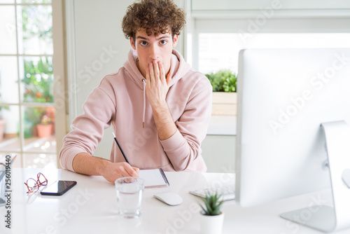 Young student man using computer and studying writing on notebook cover mouth with hand shocked with shame for mistake, expression of fear, scared in silence, secret concept