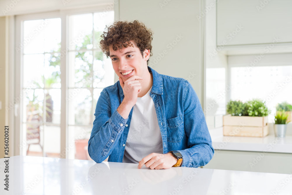 Young handsome man wearing casual denim jacket at home looking confident at the camera with smile with crossed arms and hand raised on chin. Thinking positive.