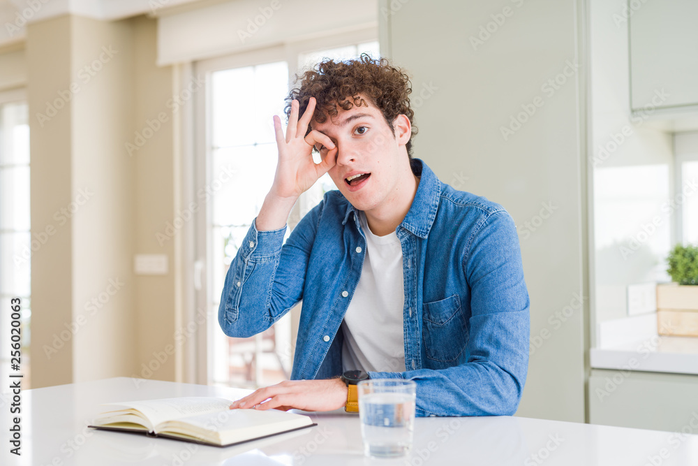Young man reading a book at home with happy face smiling doing ok sign with hand on eye looking through fingers Young man reading a book at home with happy face smiling doing ok sign with hand on eye looking through fingers
