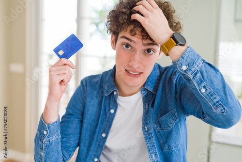 Young man holding credit card stressed with hand on head, shocked with shame and surprise face, angry and frustrated. Fear and upset for mistake.