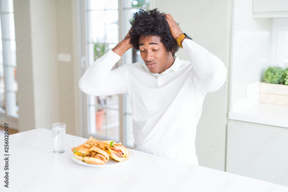 African American hungry man eating hamburger for lunch suffering from headache desperate and stressed because pain and migraine. Hands on head. African American hungry man eating hamburger for lunch suffering from headache desperate and stressed because pain and migraine. Hands on head.