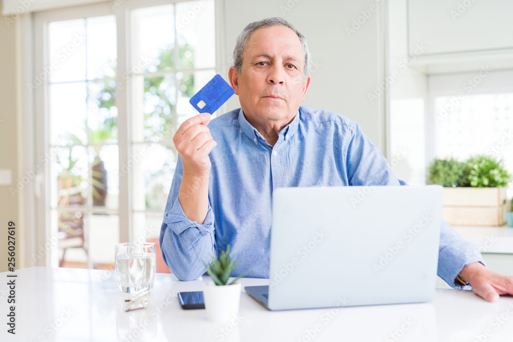 Handsome senior man shopping online using credit card and laptop at home with a confident expression on smart face thinking serious