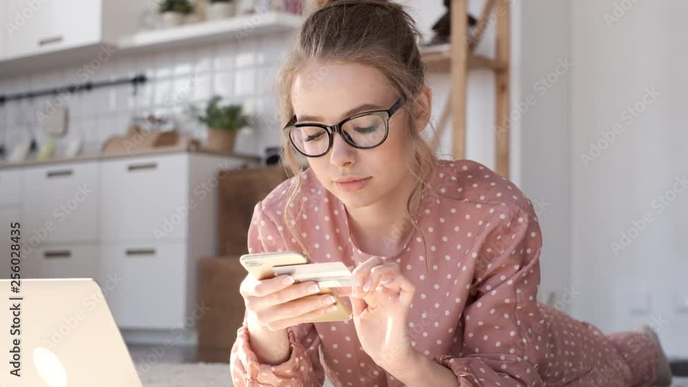 Beautiful curly blonde with glasses shopping online using laptop lying on the carpet in the living room on a sunny day. Woman shopping clothing on the internet and looking for discounts during sale. Beautiful curly blonde with glasses shopping online using laptop lying on the carpet in the living room on a sunny day. Woman shopping clothing on the internet and looking for discounts during sale.
