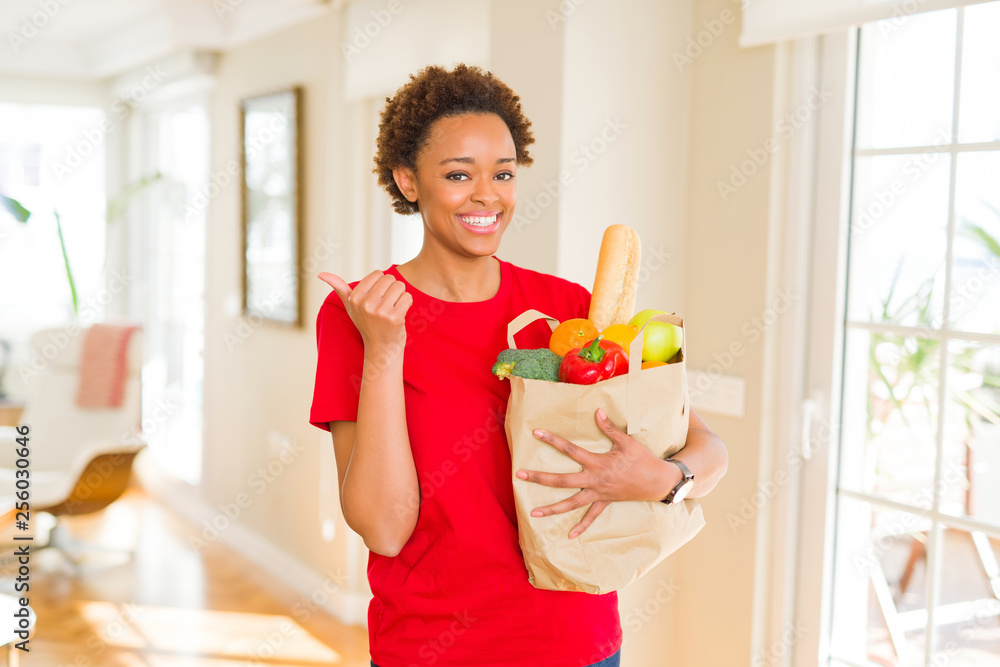 Young african american woman holding paper bag full of fresh groceries pointing and showing with thumb up to the side with happy face smiling Young african american woman holding paper bag full of fresh groceries pointing and showing with thumb up to the side with happy face smiling