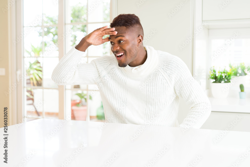Handsome african american man on white table very happy and smiling looking far away with hand over head. Searching concept.