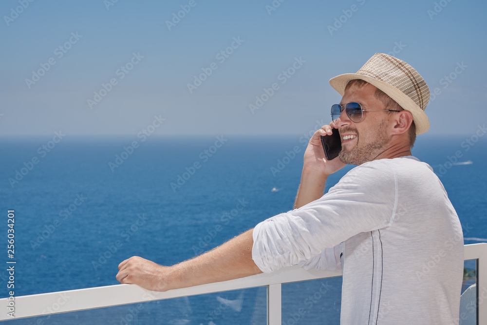European man in a sunhat and sunglasses is talking on mobile phone, while having a rest at the beautiful resort. He is sharing his emotions.