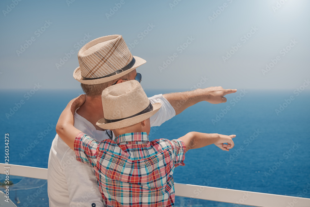 Son and dad relaxing on hotel balcony on summer holidays. They are pointing to something ahead in sea.