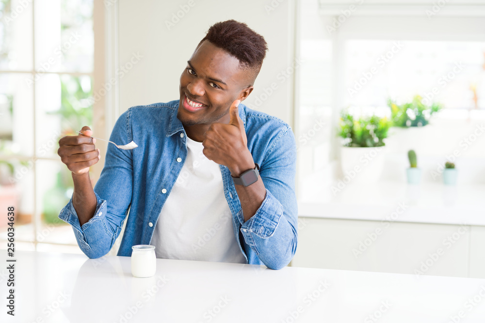 African american man eating healthy natural yogurt with a spoon happy with big smile doing ok sign, thumb up with fingers, excellent sign
