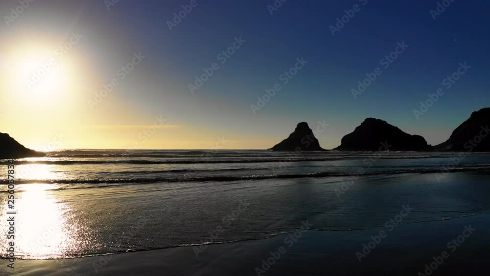 A low aerial shot flying over calm waves rolling past silhouetted rocks towards the beach along the coastline of Oregon during a summer sunset with blue sky in the background. A low aerial shot flying over calm waves rolling past silhouetted rocks towards the beach along the coastline of Oregon during a summer sunset with blue sky in the background.