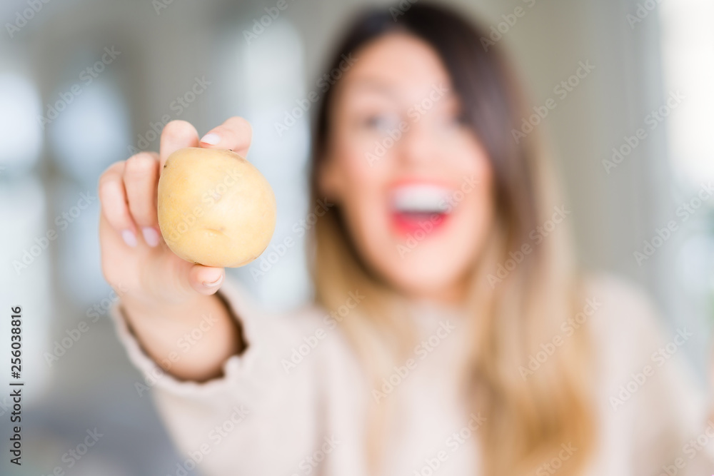 Young beautiful woman holding fresh potato at home very happy and excited, winner expression celebrating victory screaming with big smile and raised hands Young beautiful woman holding fresh potato at home very happy and excited, winner expression celebrating victory screaming with big smile and raised hands