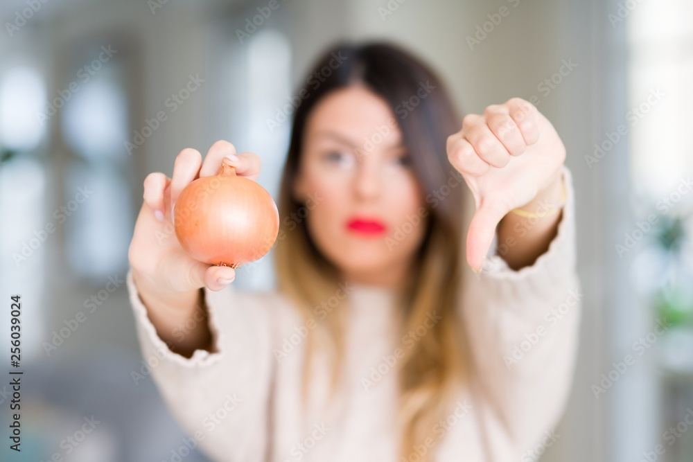 Young beautiful woman holding fresh onion at home with angry face, negative sign showing dislike with thumbs down, rejection concept Young beautiful woman holding fresh onion at home with angry face, negative sign showing dislike with thumbs down, rejection concept