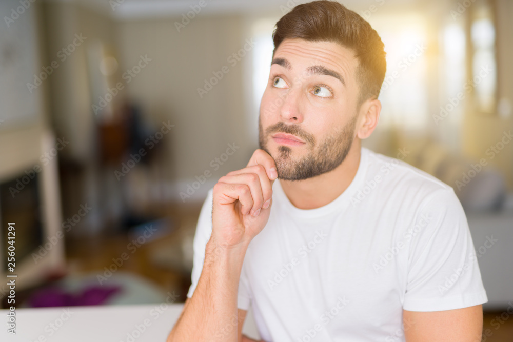 Young handsome man wearing casual white t-shirt at home with hand on chin thinking about question, pensive expression. Smiling with thoughtful face. Doubt concept. Young handsome man wearing casual white t-shirt at home with hand on chin thinking about question, pensive expression. Smiling with thoughtful face. Doubt concept.
