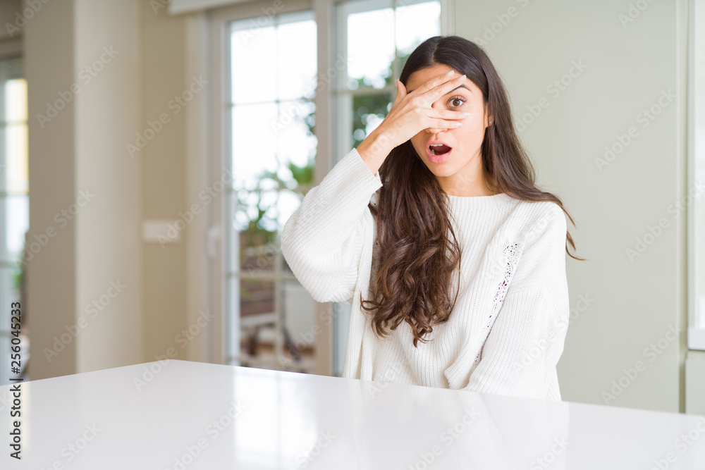 Young beautiful woman at home on white table peeking in shock covering face and eyes with hand, looking through fingers with embarrassed expression. Young beautiful woman at home on white table peeking in shock covering face and eyes with hand, looking through fingers with embarrassed expression.