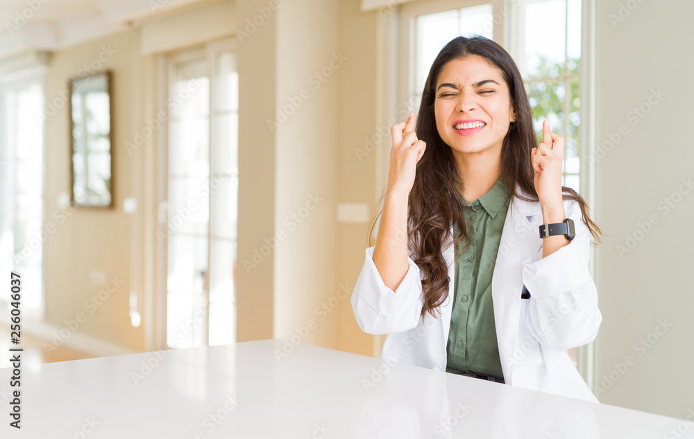 Young woman wearing medical coat at the clinic as therapist or doctor smiling crossing fingers with hope and eyes closed. Luck and superstitious concept. Young woman wearing medical coat at the clinic as therapist or doctor smiling crossing fingers with hope and eyes closed. Luck and superstitious concept.