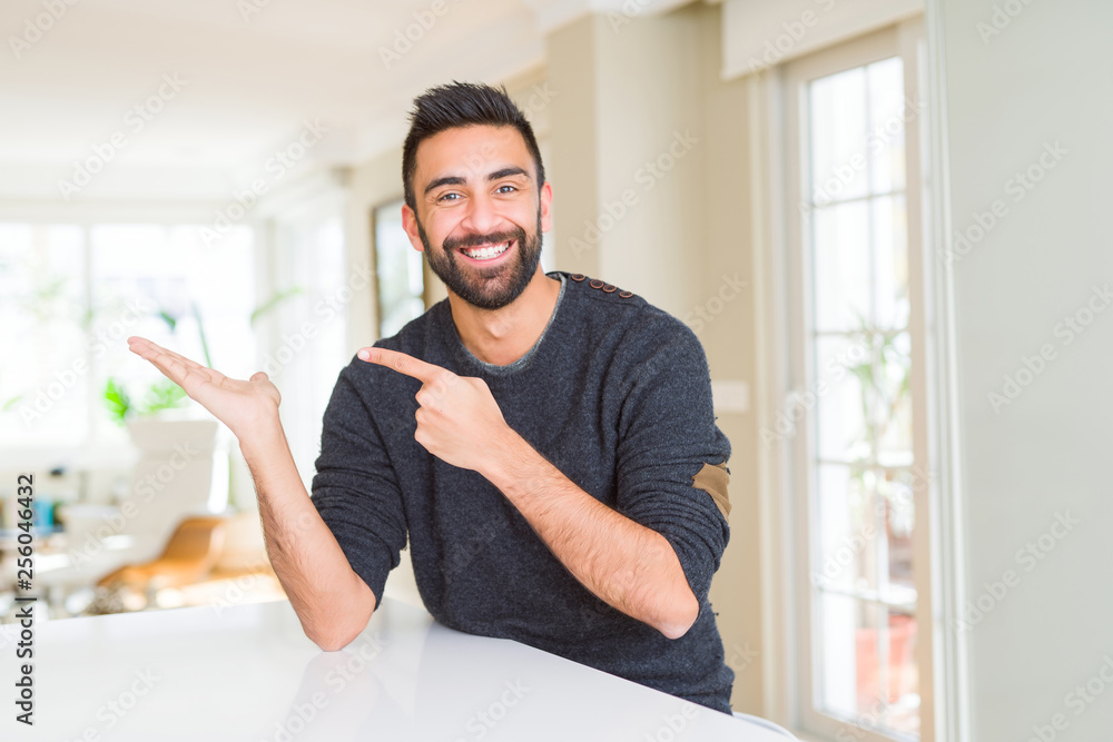 Handsome hispanic man wearing casual sweater at home amazed and smiling to the camera while presenting with hand and pointing with finger. Handsome hispanic man wearing casual sweater at home amazed and smiling to the camera while presenting with hand and pointing with finger.