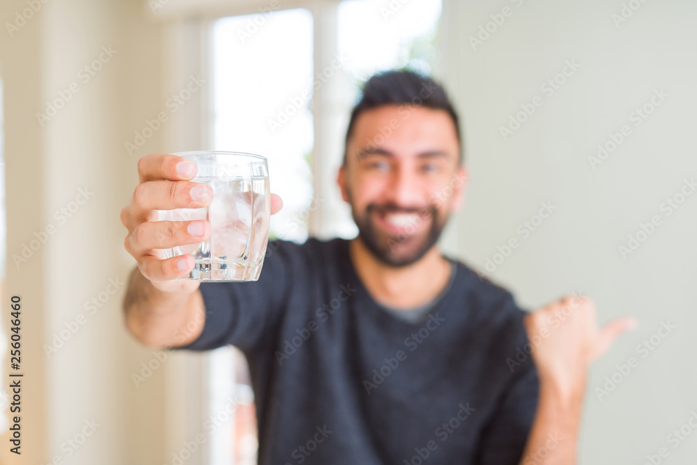 Handsome hispanic man drinking a fresh glass of water pointing and showing with thumb up to the side with happy face smiling Handsome hispanic man drinking a fresh glass of water pointing and showing with thumb up to the side with happy face smiling