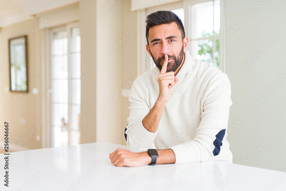 Handsome hispanic man wearing casual white sweater at home asking to be quiet with finger on lips. Silence and secret concept.