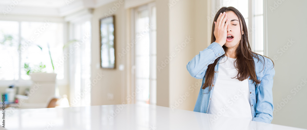 Wide angle picture of beautiful young woman sitting on white table at home Yawning tired covering half face, eye and mouth with hand. Face hurts in pain.