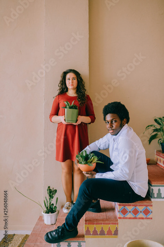 Interracial lifestyle couple portrait. Dark-skinned african man siiting on stairs with plant in pot at hands. Adult woman with home flower in hadns standing behind black nigerian male. Loving family. photo