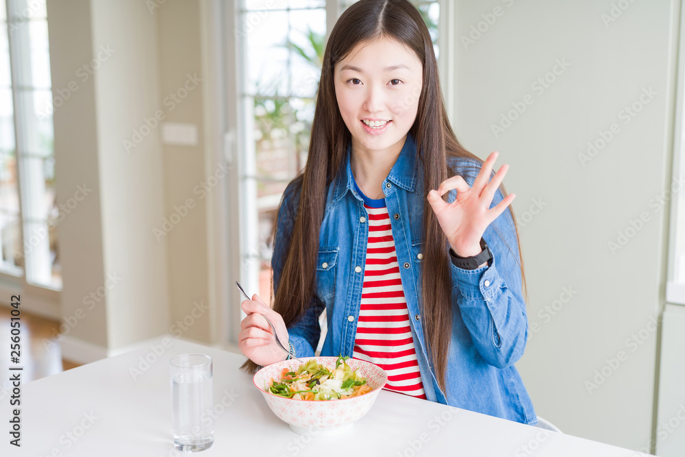 Beautiful Asian woman eating healthy pasta salad doing ok sign with fingers, excellent symbol Beautiful Asian woman eating healthy pasta salad doing ok sign with fingers, excellent symbol