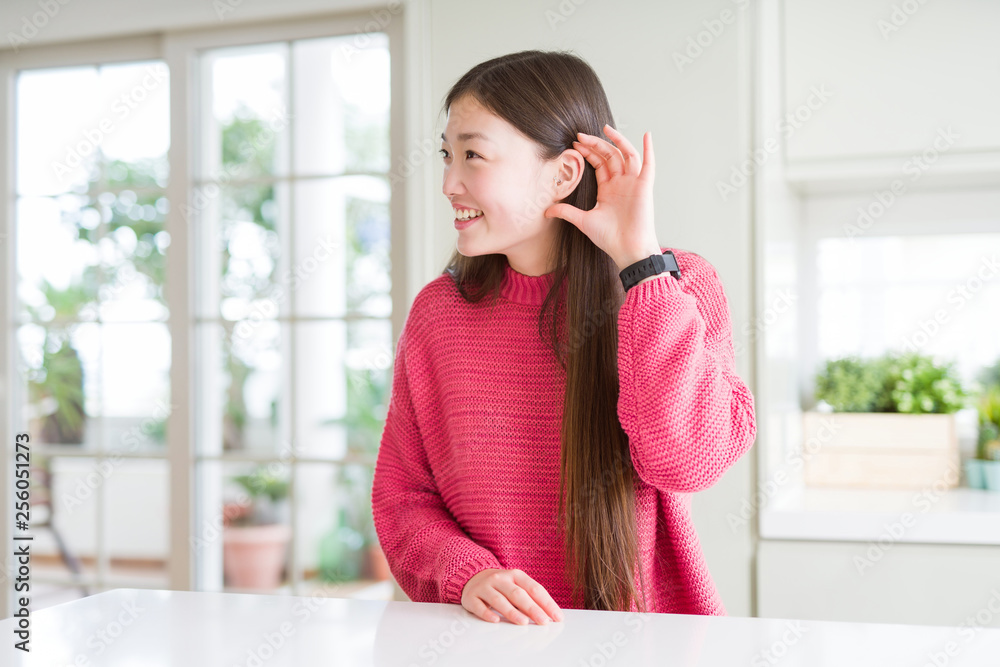Beautiful Asian woman wearing pink sweater on white table smiling with hand over ear listening an hearing to rumor or gossip. Deafness concept. Beautiful Asian woman wearing pink sweater on white table smiling with hand over ear listening an hearing to rumor or gossip. Deafness concept.