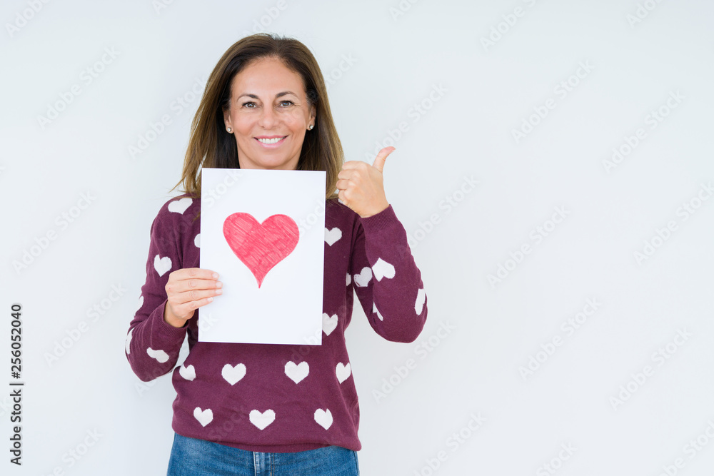 Middle age woman holding card gift with red heart over isolated background pointing and showing with thumb up to the side with happy face smiling Middle age woman holding card gift with red heart over isolated background pointing and showing with thumb up to the side with happy face smiling